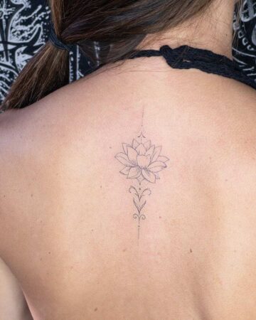 Elegant lotus flower tattoo on a woman's upper back, centered along her spine. The delicate black ink design features a detailed lotus blossom with ornamental flourishes above and below. A beautiful example of lotus flower tattoo art.
