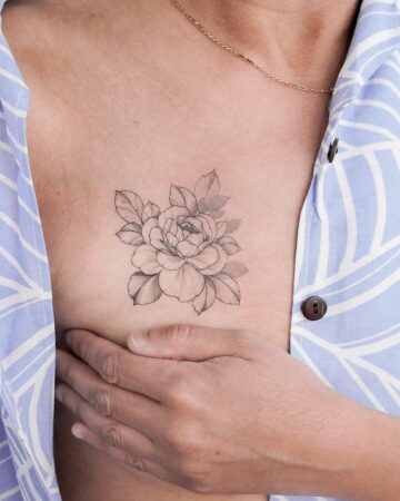 Elegant peony tattoo on a woman's chest, visible beneath a partially unbuttoned blue patterned shirt. A delicate gold chain necklace adds a subtle accent.