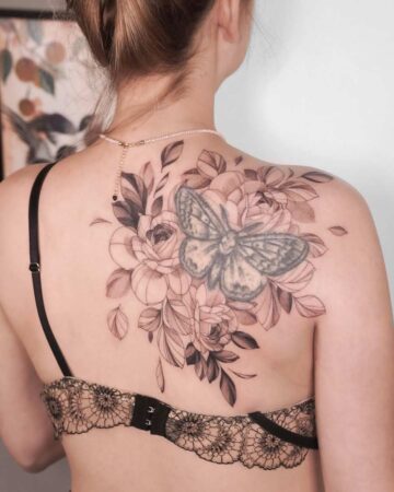 Back view of a woman with a large peony tattoo on her upper back, featuring a butterfly and detailed floral design in shades of gray and black. The tattoo extends across her shoulder blade.