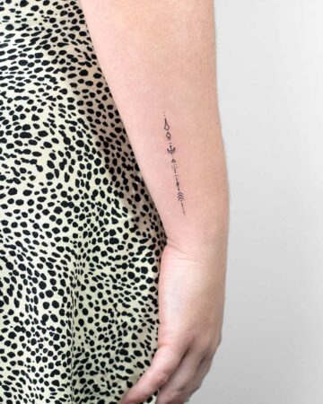 A delicate, vertical spiritual tattoo comprised of geometric symbols runs along the inner forearm. This minimalist lettering tattoo is set against a background of a black and white patterned dress. Created by our tattoo artists in Canggu, Bali