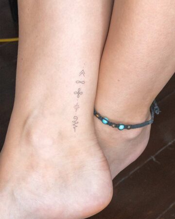 A delicate ankle tattoo featuring a vertical stack of spiritual symbols is seen on a fair-skinned leg, paired with a beaded anklet on the other ankle. This spiritual tattoo gallery showcases unique designs. Created by our tattoo artists in Canggu, Bali
