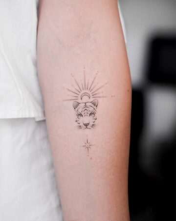 Close-up of a forearm tattoo featuring a minimalist tiger face design with radiating lines above resembling a sun or halo, and a star below. A subtle example of tiger Tattoos.
