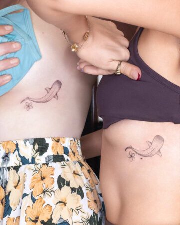 Two women displaying matching side tattoos of a fish and a frangipani flower. A delicate Frangipani Plumeria & Hibiscus Tattoo design, subtly placed on their skin.