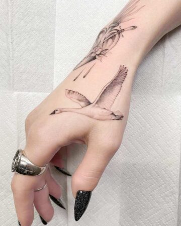 A hand featuring a stunning micro realism tattoo of a swan in flight, with a second tattoo of a heart and flower on the wrist. The hand is adorned with a bold silver ring and dark, pointed nails. Done by our expert tattoo artists at our fine line tattoo studio in Canggu, Bali.