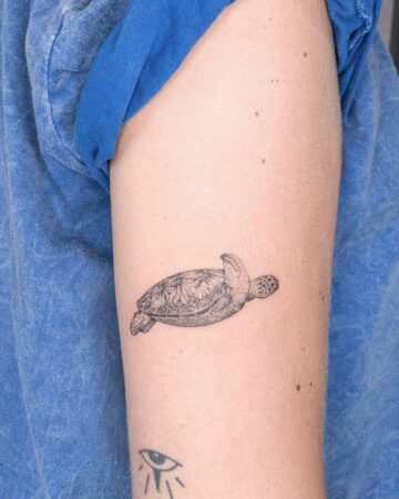 A detailed black ink tattoo of a sea turtle graces the upper arm of a person wearing a blue shirt. This aquatic creature tattoo showcases intricate shell patterns and a lifelike depiction of marine life. Created by our tattoo artists in Canggu, Bali