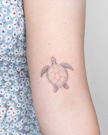 Delicate line art marine life tattoo of a sea turtle on a woman's arm. The turtle's shell is intricately detailed, set against a background of a blue floral dress.