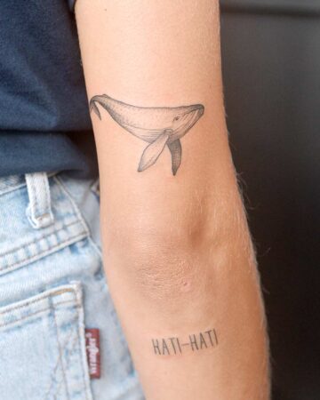 Close-up of a delicate whale tattoo on a fair-skinned arm, positioned above the elbow. Below, the words "HATI-HATI" are tattooed. The arm is set against a backdrop of denim jeans and a dark wall. Created by our tattoo artists in Canggu, Bali