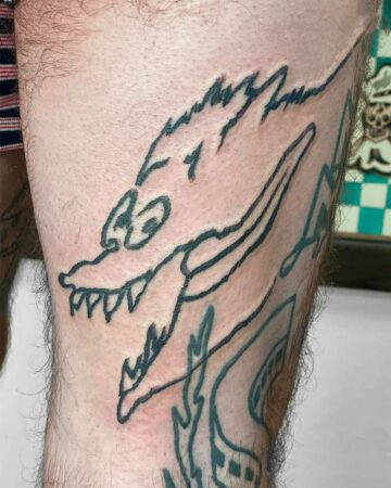 A bold, black line art dog tattoo on a hairy leg, featuring a stylized wolf-like creature with sharp teeth. A unique dog tattoo design. Created by a skilled Bali tattoo artist in Canggu.