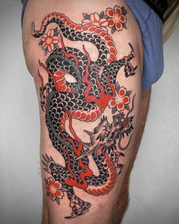 A bold Japanese dragon tattoo in black, red, and gray scales coils around a thigh, accented with cherry blossoms. A fine example of Japan tattoo art. Created in a Bali tattoo studio.