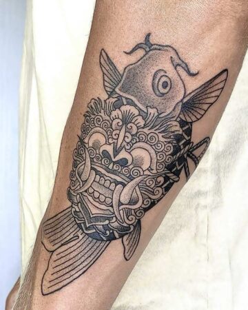 Forearm tattoo featuring a Barong mask integrated with a koi fish design in black ink. A unique Barong tattoo displaying Balinese art. Created by the best expert tattoo artists in Canggu Bali for unique tattoos.