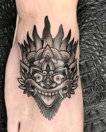 A striking black and grey Barong tattoo on a foot. The tattoo features a detailed Barong mask design with sharp lines and shading. Created by the best expert tattoo artists in Canggu Bali.
