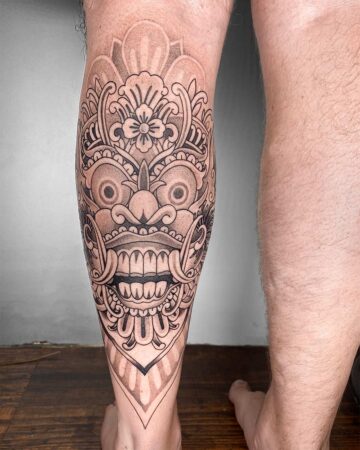Detailed black and grey Barong tattoo on a man's calf, showcasing intricate Balinese mask art. A striking example from our Barong tattoo gallery. Created by the best expert tattoo artists in Canggu Bali.