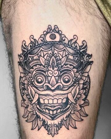 Close-up of a detailed black and gray Barong tattoo on a person's thigh. The intricate design features the iconic mask with bulging eyes, sharp teeth, and ornate crown. A striking example of Barong tattoo art in Bali. Created by the best expert tattoo artists in Canggu Bali for your next Bali tattoo.