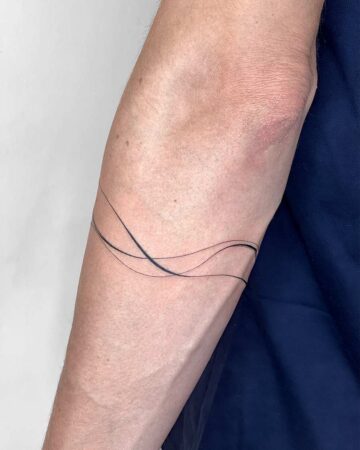 Minimalist bracelet tattoo design on a fair-skinned arm. Thin, black lines create a delicate, abstract bracelet around the forearm. Created by the best expert tattoo artists in Canggu Bali.