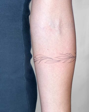 Close-up of a delicate, fine-line bracelet tattoo of leaves and vines encircling a fair-skinned arm. A simple and elegant body art design. Created by the best tattoo artists in Canggu Bali for fine line tattoos.