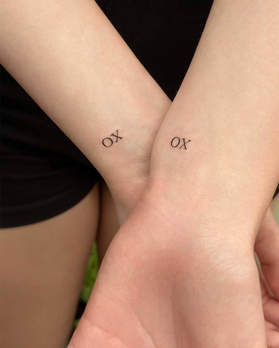 A minimalist couple's tattoo on the inner wrists. Each person has the letters "XO" tattooed in a clean, serif typography, symbolizing hugs and kisses.