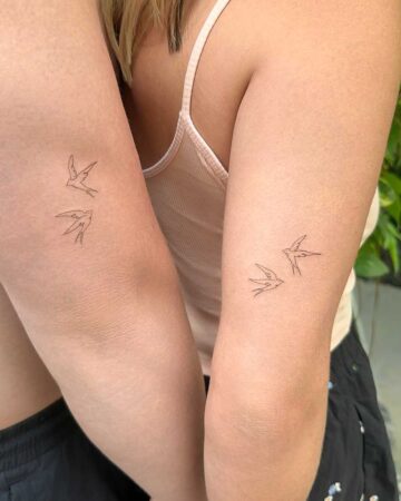 Matching bird tattoos on two forearms. Each tattoo features two small swallows in flight, created with a single-line, minimalist drawing style.