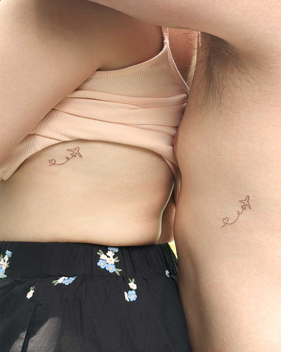 Two people standing side-by-side showing matching tattoos on their ribs and torso. The design is a tiny paper airplane leaving a heart-shaped smoke trail behind it.