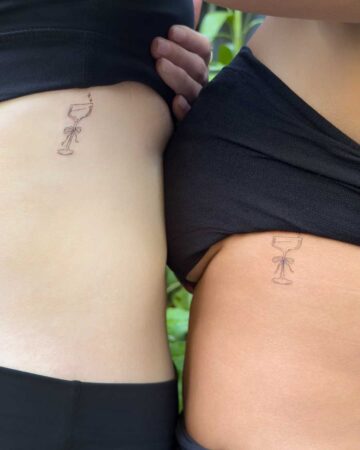 Two friends show off their new matching fine line tattoos on their ribcages. The design is a minimalist martini glass with a small bow around the stem and a pair of tiny star sparkles. Their skin tones are different, highlighting the universal appeal of the delicate black ink work.