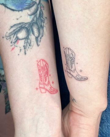 A close-up of two forearms featuring matching cowboy boot tattoos in a minimalist fine line style. The boot on the left is rendered in vibrant red ink, while the boot on the right is in classic black ink. Both designs include intricate decorative stitching and are surrounded by tiny, delicate sparkles.