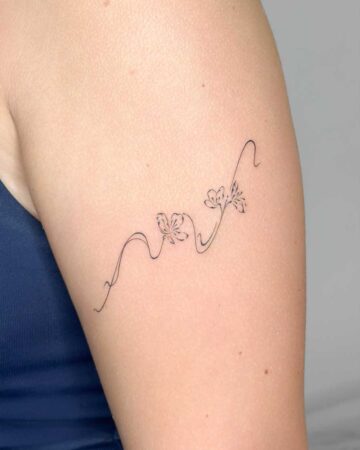 Delicate black ink floral tattoo on an arm, featuring a winding vine with two small, minimalist flowers. This subtle floral tattoo is a beautiful example of fine line artistry. Created in the multi style tattoo studio in Canggu Bali, Bali tattoo studio, best tattoo Canggu.