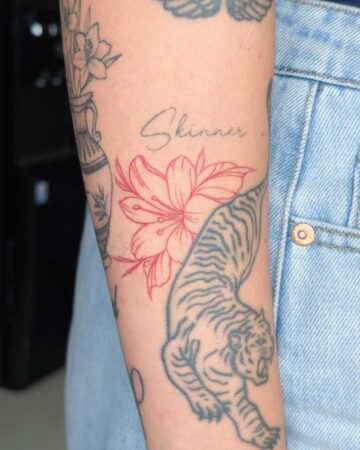 Close-up of a person's arm showcasing a delicate pink floral tattoo with the name "Skinner" in script above it, alongside a grey tiger tattoo. This floral tattoo art is a beautiful example of tattoo studio work in Canggu Bali. Created in the multi style tattoo studio in Canggu Bali, Bali tattoo.