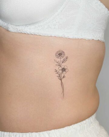 Delicate black ink floral tattoo on a woman's ribcage, featuring two stylized flowers and sprigs of greenery. This fine line floral tattoo showcases minimalist botanical art. Created in the multi style tattoo studio in Canggu Bali, Bali tattoo studio, best tattoo Canggu.