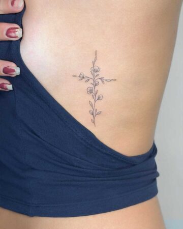 Delicate floral cross tattoo on a person's ribcage, showcasing a minimalist line art style with small blossoms and leaves. This is a beautiful example of a floral tattoo, created in the multi style tattoo studio in Canggu Bali.