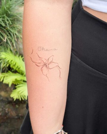 Delicate floral tattoo of a plumeria flower with the word "Ohana" in script on a person's arm, set against a blurred green leafy background. Created in the multi style tattoo studio in Canggu Bali.