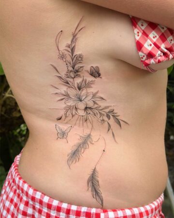 A delicate floral tattoo adorns a person's ribcage, featuring intricate flowers, leaves, and butterflies. This beautiful floral tattoo design incorporates feathers, creating a dreamcatcher-like effect. Created in the multi style tattoo studio in Canggu Bali, a top Bali tattoo destination.