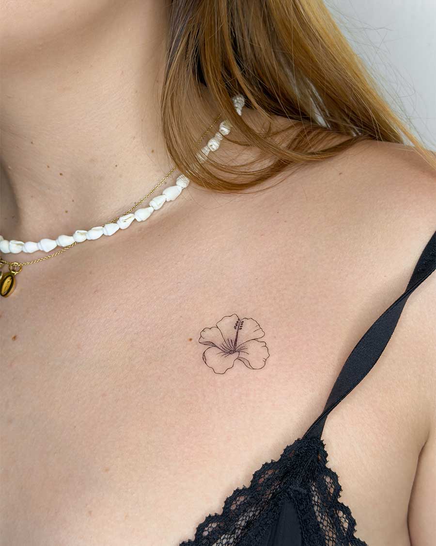 Delicate black ink hibiscus tattoo on a woman's collarbone, a subtle floral design. Bali tattoo studio inspiration.