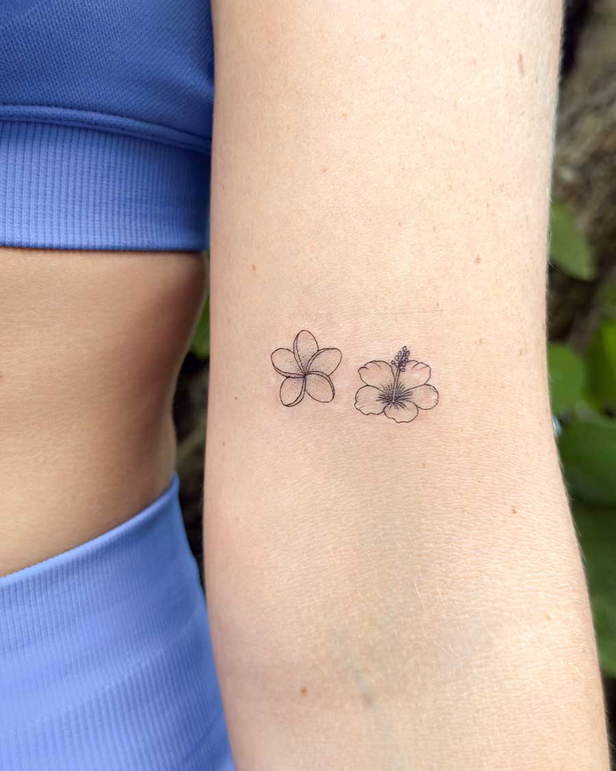 Delicate black ink line drawings of a frangipani and a hibiscus flower adorn a person's arm, showcasing minimalist floral tattoo art. Created in the multi style tattoo studio in Canggu Bali.