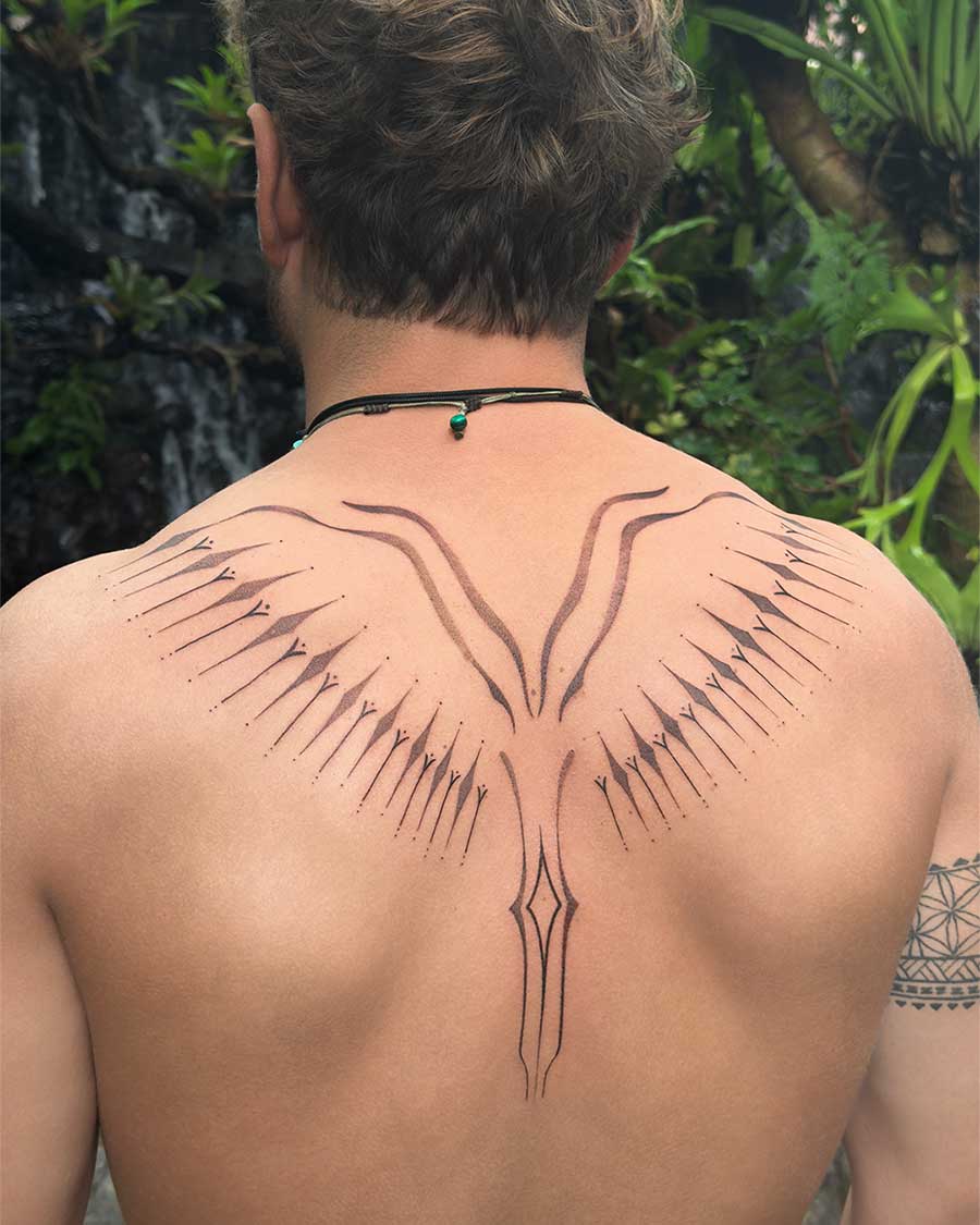 Back view of a man with an ornamental tattoo resembling stylized wings on his upper back. The intricate black ink design features delicate lines and dots, set against a lush green natural background with a waterfall. Created in the multi style tattoo studio in Canggu Bali, Bali tattoo studio, Bali ornamental tattoo.